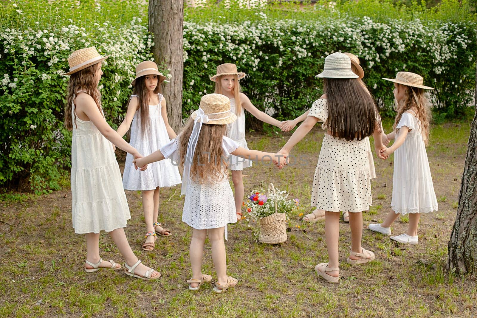 Group of preteen girls wearing white light dresses and wicker hats holding hands and dancing in circle on lawn in green summer park. Happy childhood concept