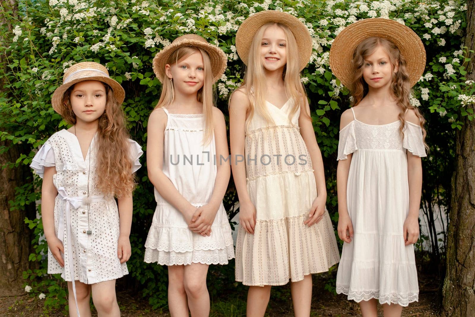 Smiling tween girls wearing light dresses and straw hats standing in summer park on background of green bush blooming with white flowers, cropped shot