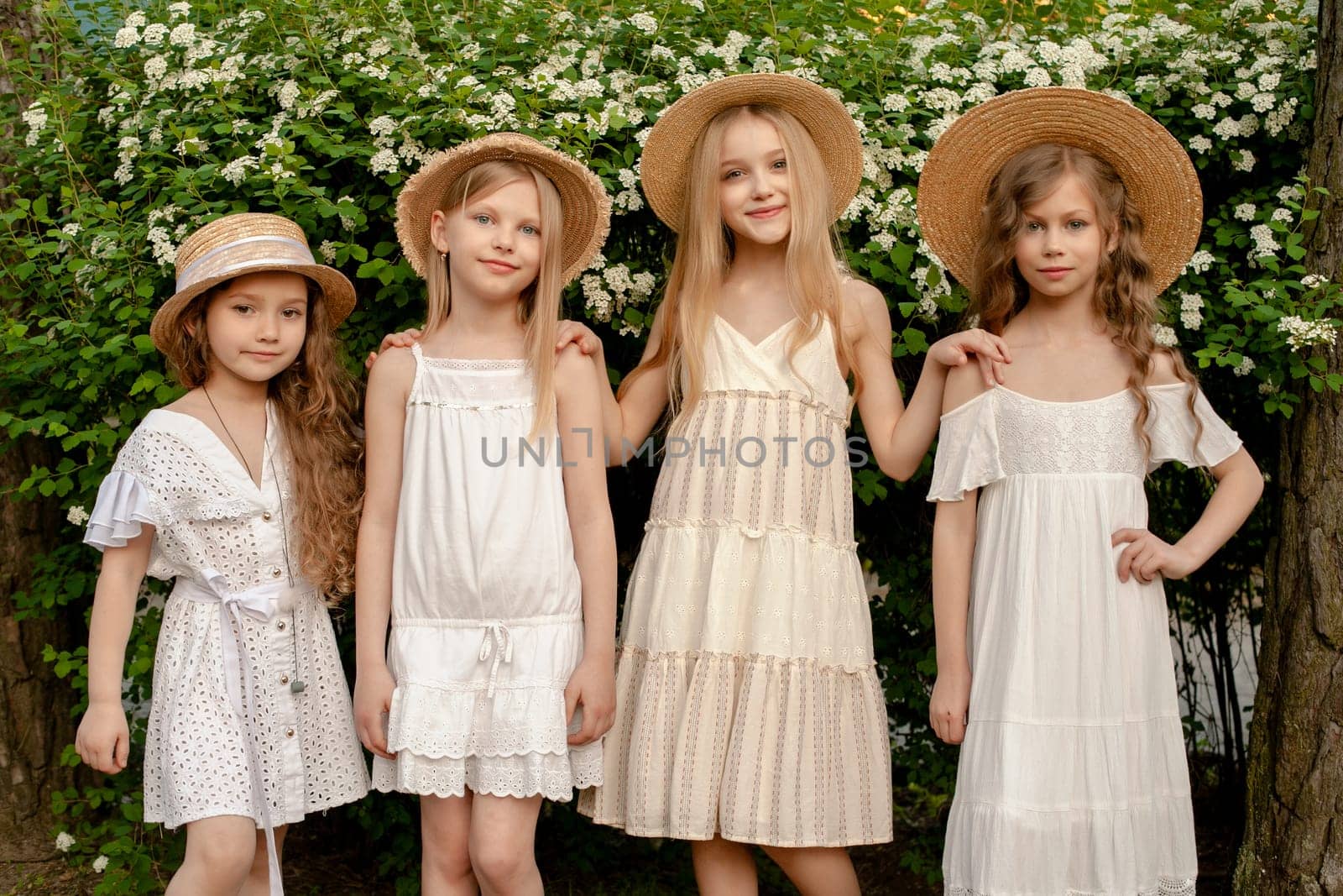 Group portrait of friendly happy preteen girls in country style dresses and wicker staw hats standing outdoors on background of large green shrub with white flowers on summer day