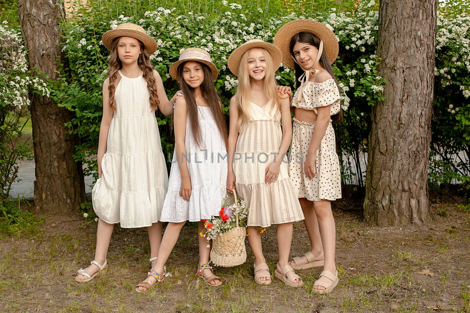 Full-length portrait of smiling cute stylish preteen girls wearing light dresses and wicker sun hats standing near green blooming bush in park on summer day, holding basket with flowers