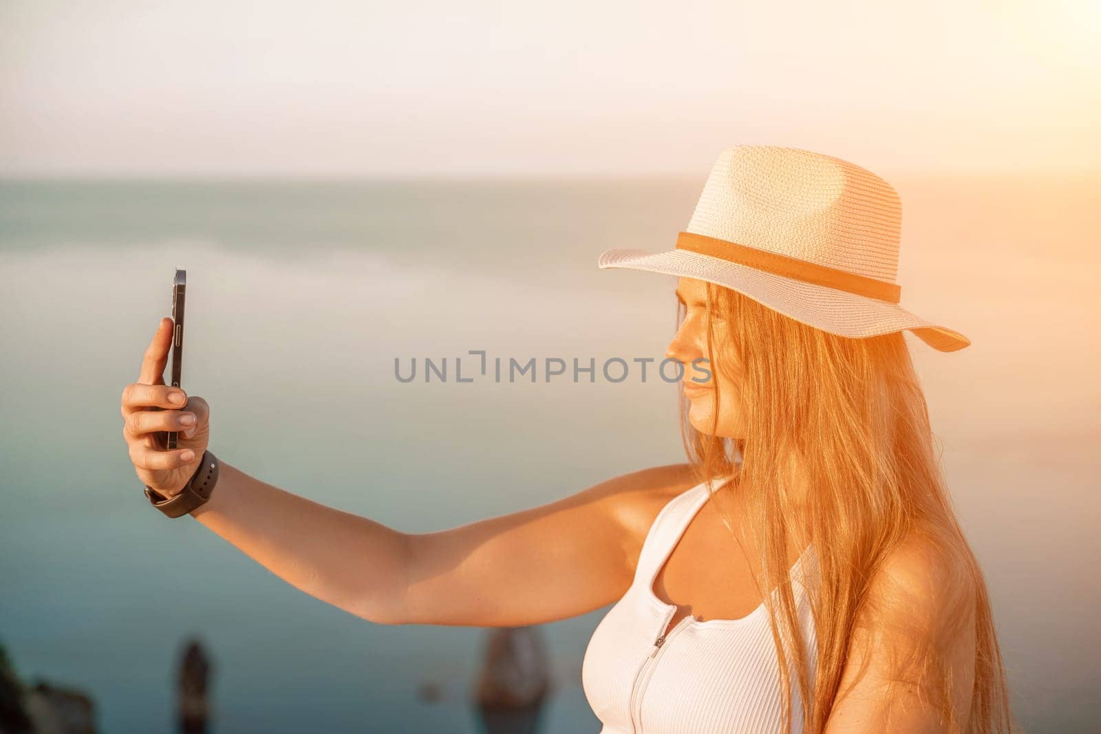 Selfie woman in a hat, white tank top, and shorts captures a selfie shot with her mobile phone against the backdrop of a serene beach and blue sea