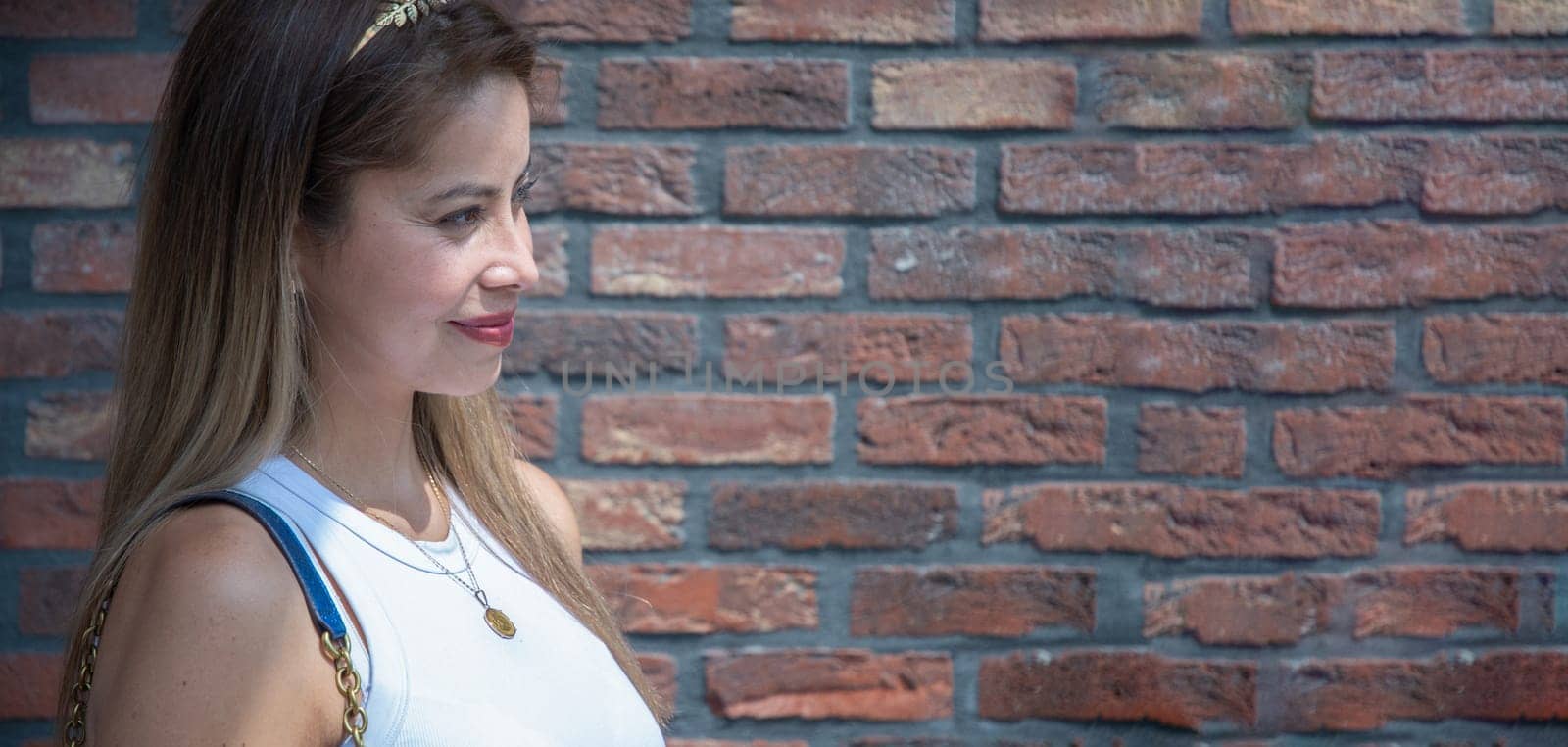 Young blonde woman in stylish clothes looks away smiling near the red brick wall of the house on the street on a bright sunny day, light and shadow, banner with Copy Space, High quality photo