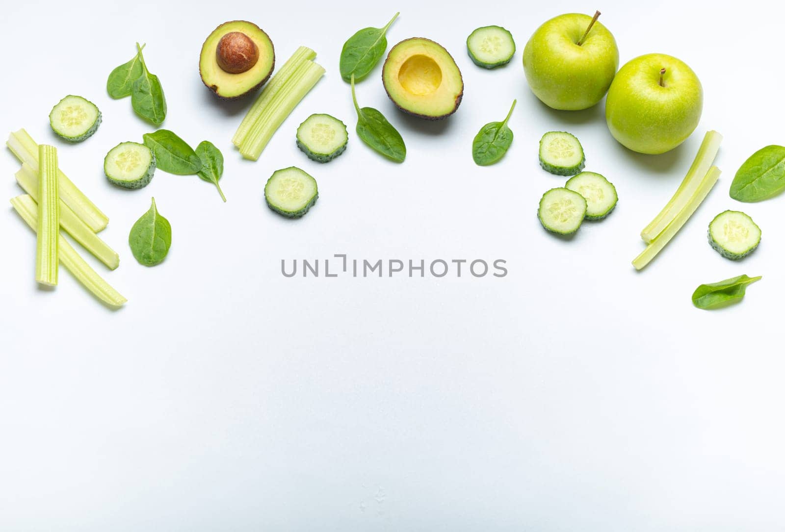 Healthy food border with green fresh vegetables and fruit on simple white background: apples, spinach, celery sticks, spinach leaves, avocado, cucumber. Space for text by its_al_dente