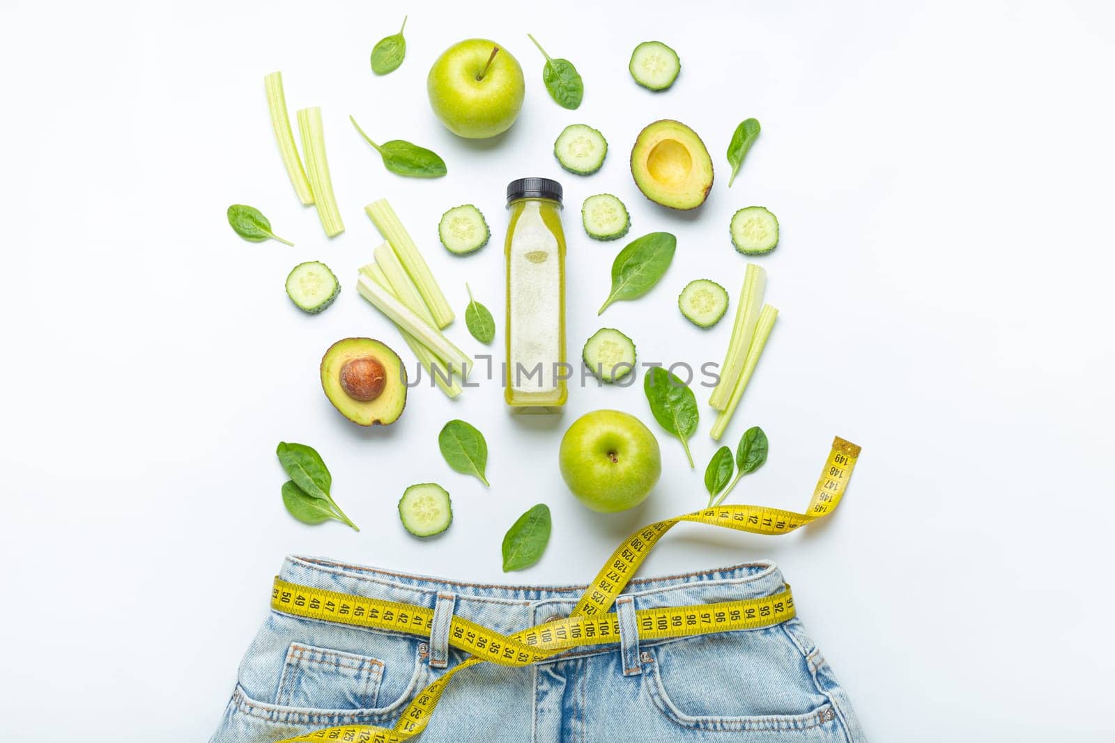 Green fruit, vegetables, smoothie falling into jeans and yellow measuring tape instead of belt on white background. Concept of healthy food for weight loss, detox, diet, healthy clean nutrition