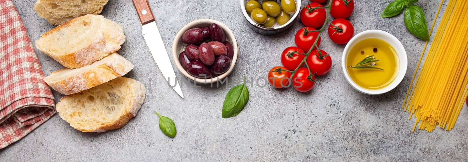 Food composition with sliced ciabatta, olives, olive oil, spaghetti, fresh basil, cherry tomatoes on gray concrete stone rustic background top view, copy space. Italian cuisine concept