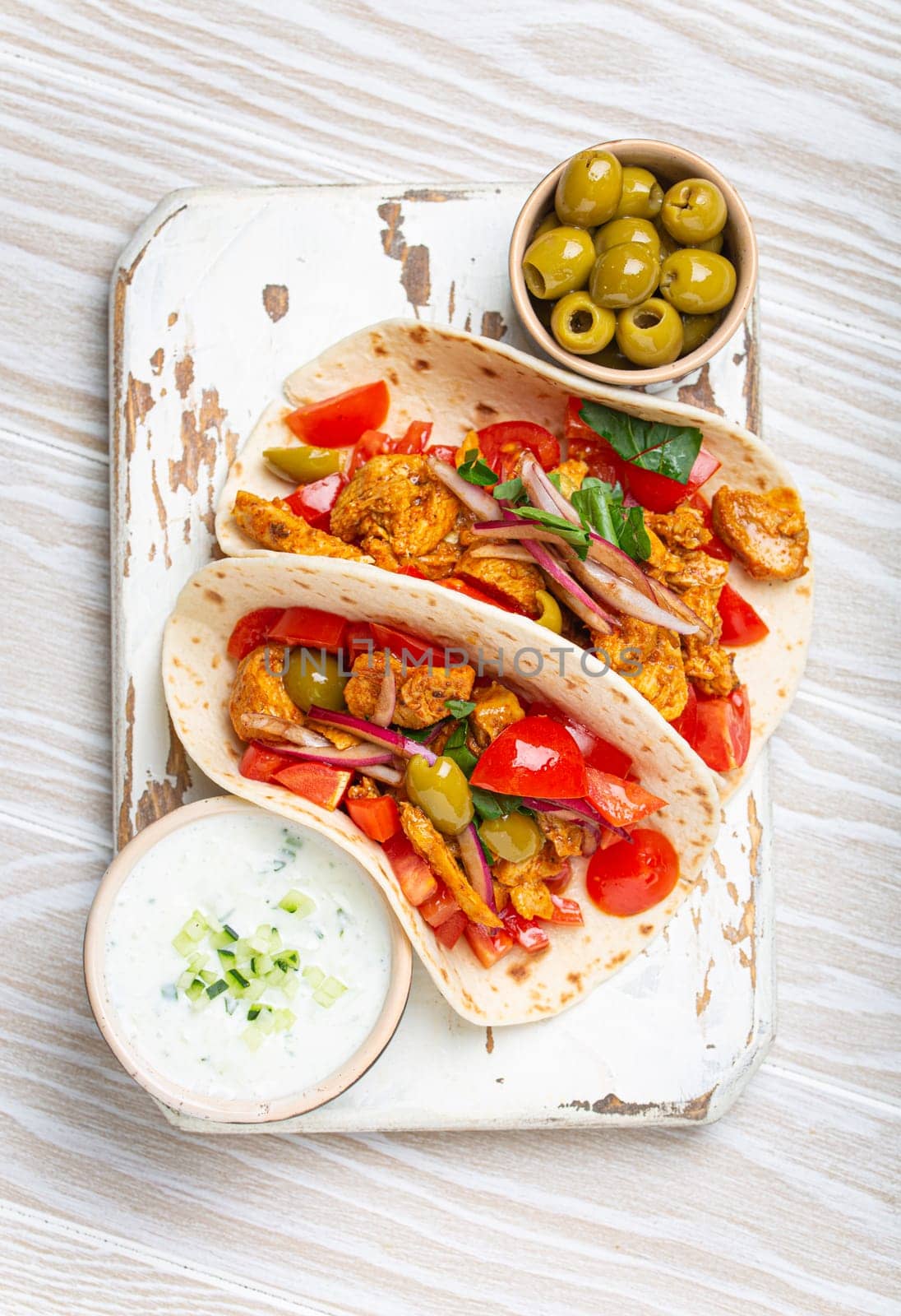 Traditional Greek Dish Gyros: Pita bread Wraps with vegetables, meat, herbs, olives on rustic wooden cutting board with Tzatziki sauce, olive oil top view, white wooden summer background.