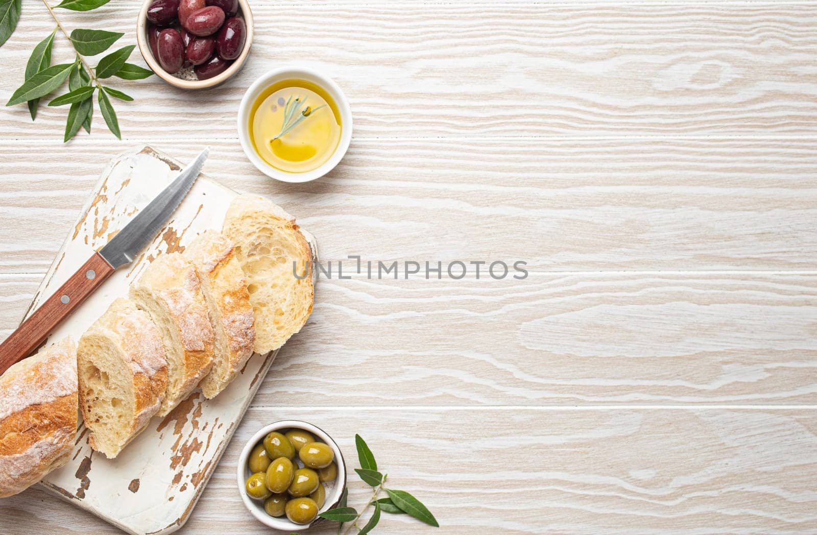 Sliced fresh ciabatta on cutting kitchen board, green and brown olives, olive oil with rosemary, olive tree branches on white wooden rustic background top view by its_al_dente
