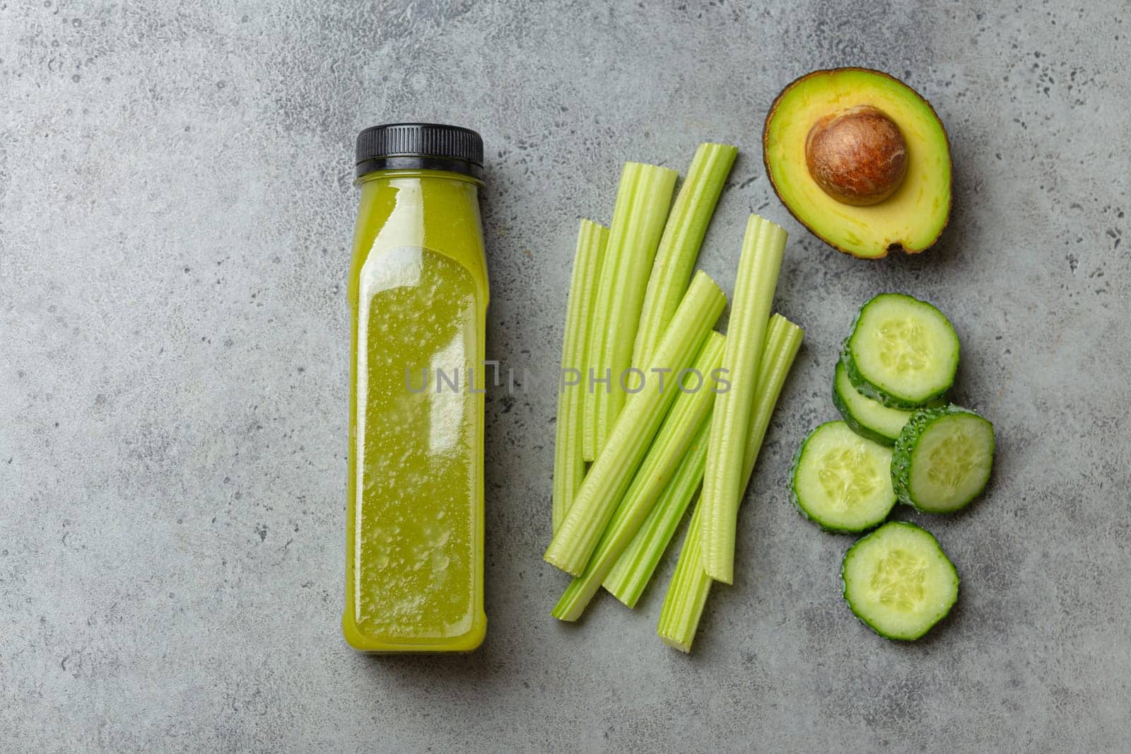 Bottle of green vegetable smoothie with fresh celery sticks, cut avocado, cucumber on gray concrete background top view. Diet beverage, healthy nutrition, detox concept by its_al_dente