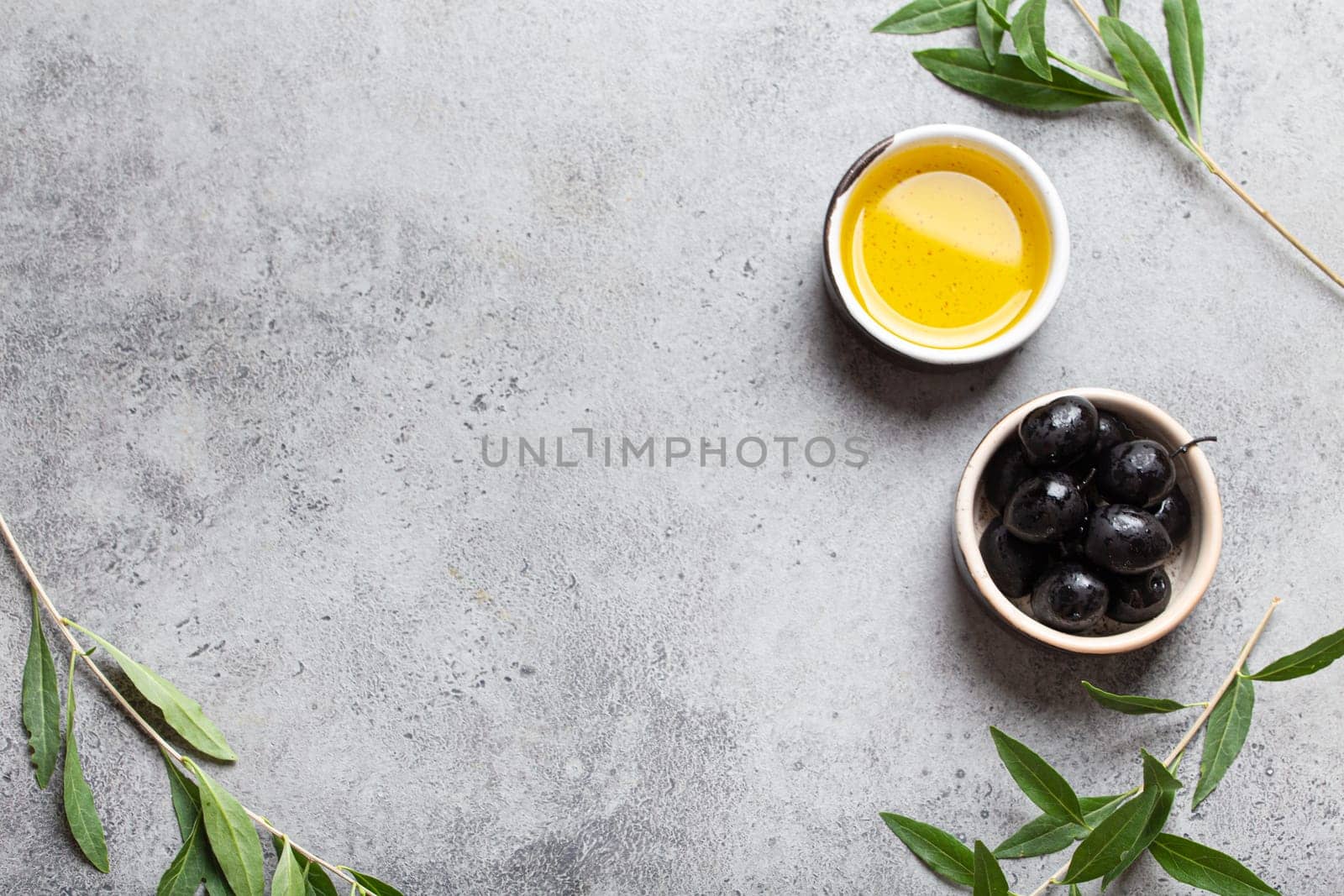 Black olives and extra virgin olive oil in little bowl with olive tree branches top view on grey concrete stone background, space for text by its_al_dente