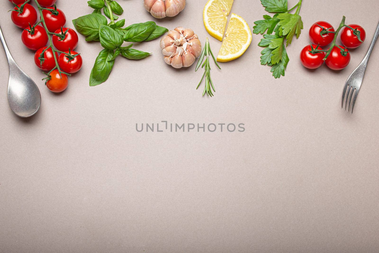 Composition with branch of fresh cherry tomatoes, herbs, garlic cloves, lemon wedges, kitchen spoon and fork on minimalistic grey clean background, overhead shot with copy space by its_al_dente