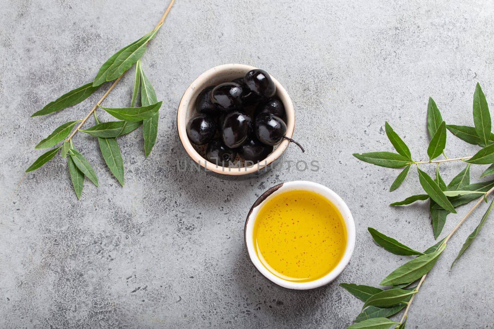 Black olives and extra virgin olive oil in little bowl with olive tree branches top view on grey concrete stone background by its_al_dente