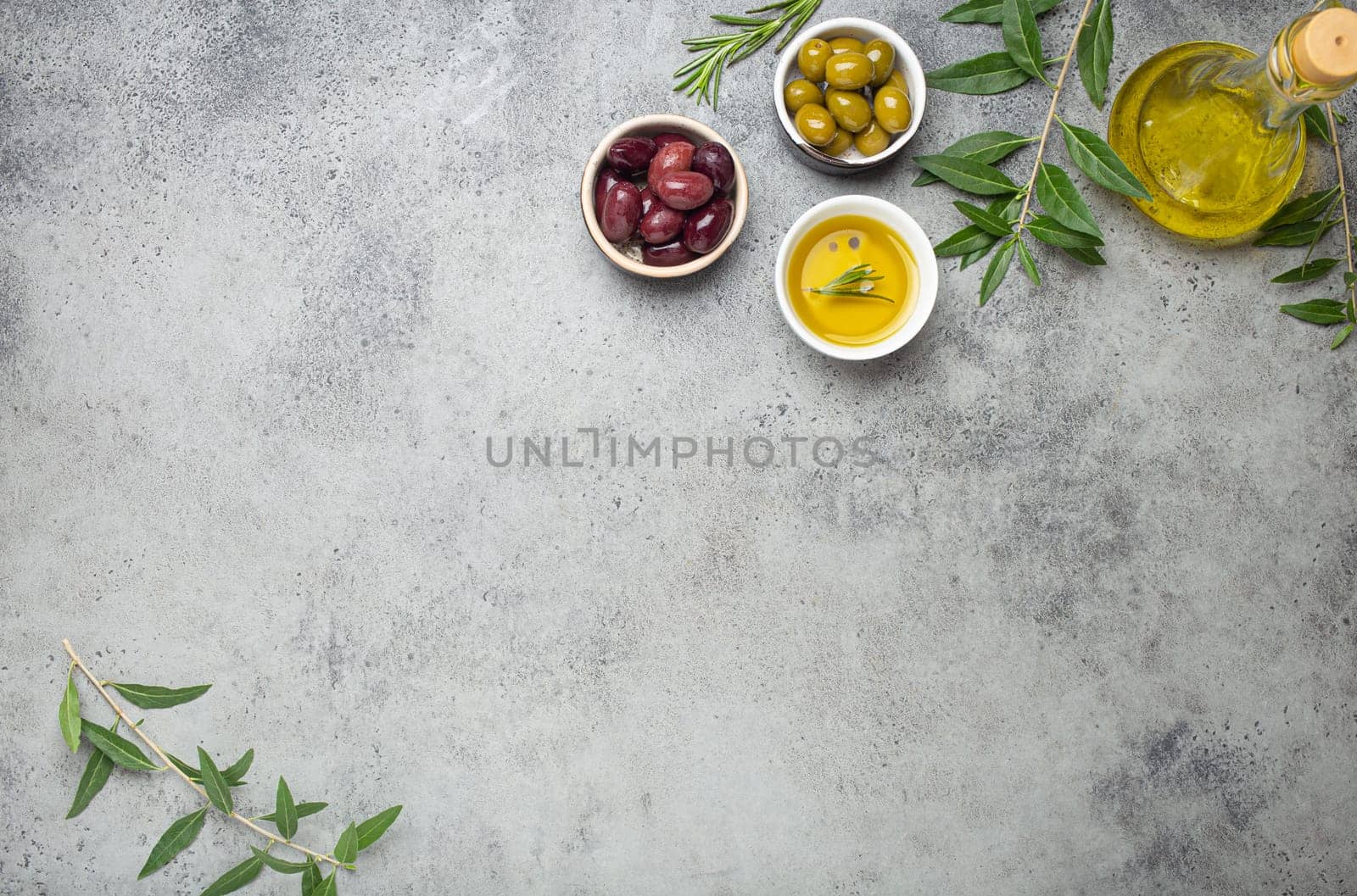 Composition with green and brown olives, extra virgin olive oil in glass bottle, olive tree branches on gray concrete stone rustic background overhead, copy space border
