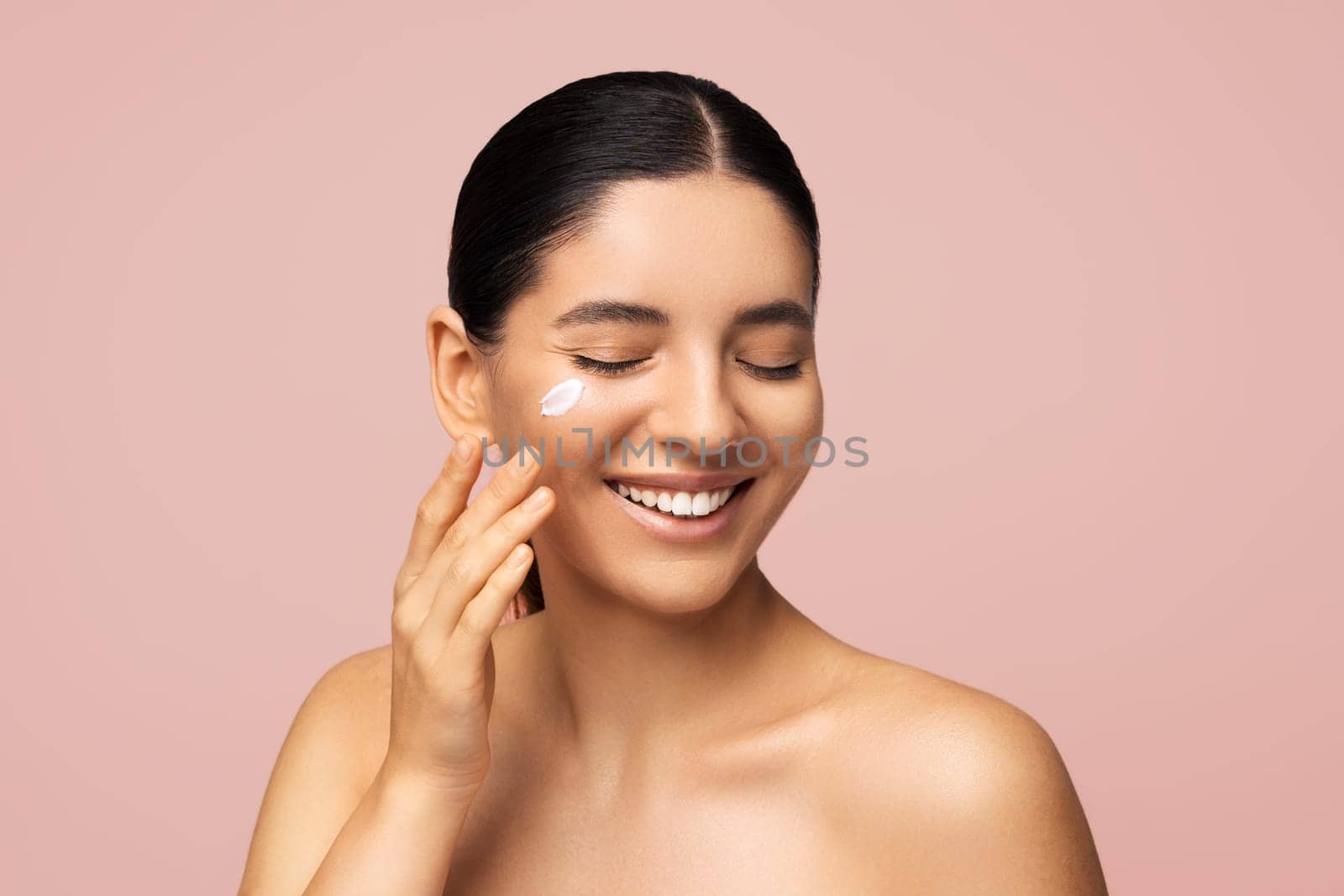 Beauty portrait. Attractive multi-ethnic woman 25s with cheerful smile applying cream on face for skincare.