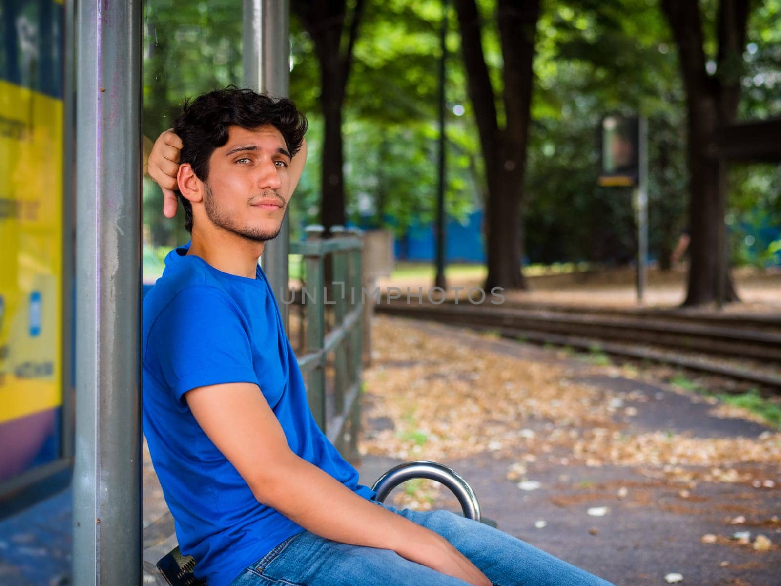 Three quarters shot of one handsome young man in urban setting waiting tram at bus stop, wearing blue polo shirt