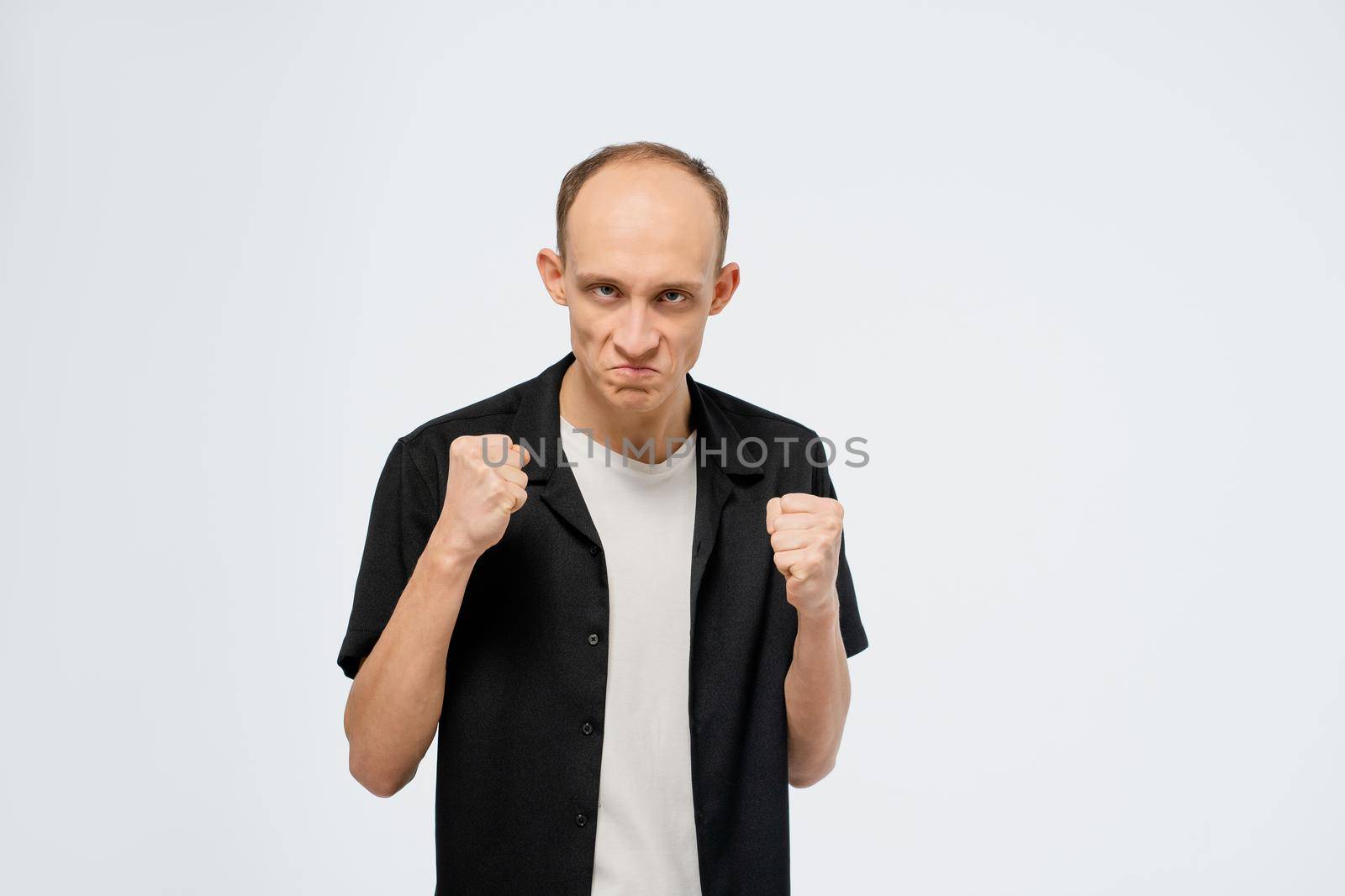 Defending himself stands in a stance with ready fists young bald man in black shirt with white t-shirt under. Angry young adult man showing he is ready for fight isolated on white background by LipikStockMedia