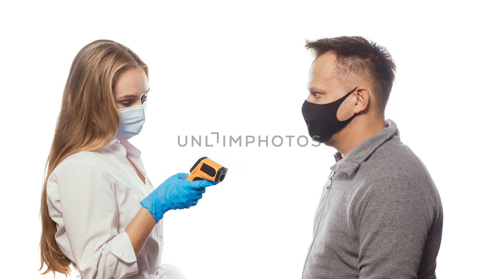 Looking at non-contact thermometer nurse testing employee's or patients body temperature wearing a medical face mask. Sad man wearing reusable face mask isolated on white background by LipikStockMedia