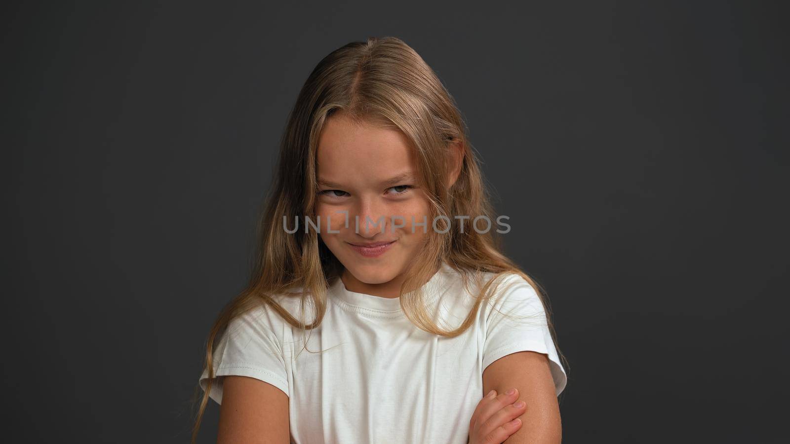 Close up. Little girl of 8,10 years with hands folded looks looks questioningly at the camera wearing white t shirt isolated on dark grey or black background.