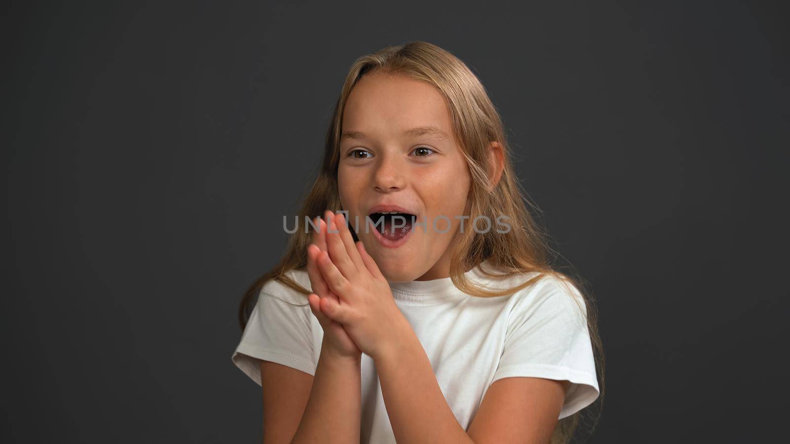 Excited little girl standing with hands put together happily looking a side of camera wearing white t-shirt isolated on dark grey or black background by LipikStockMedia