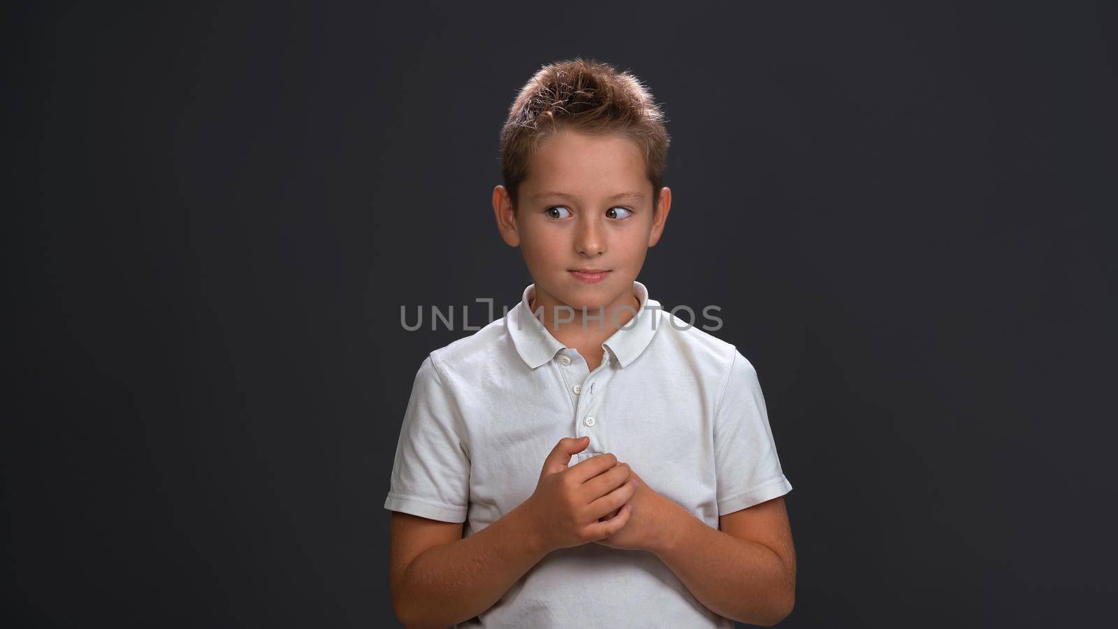 Frustrated boy in white shirt holding hands together in front of him while looking sideways at upper corner. Isolated on black background by LipikStockMedia