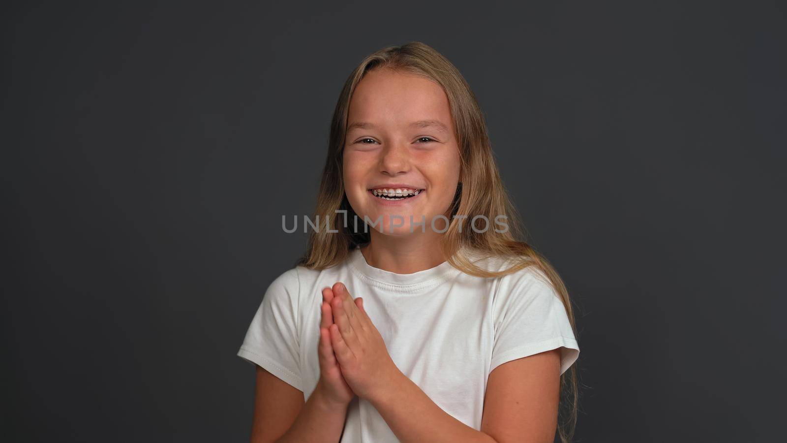 Happy charming little girl with hands put together looking a side of camera, wearing white t-shirt isolated on dark grey or black background by LipikStockMedia