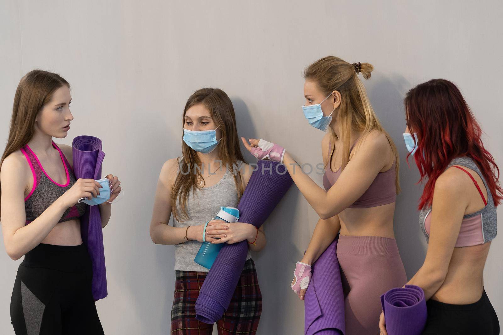 Convincing friend to wear medical face protective mask athletic girls in sports out fits standing next to the wall holding a yoga mat, talking to each other, standing at a grey background by LipikStockMedia