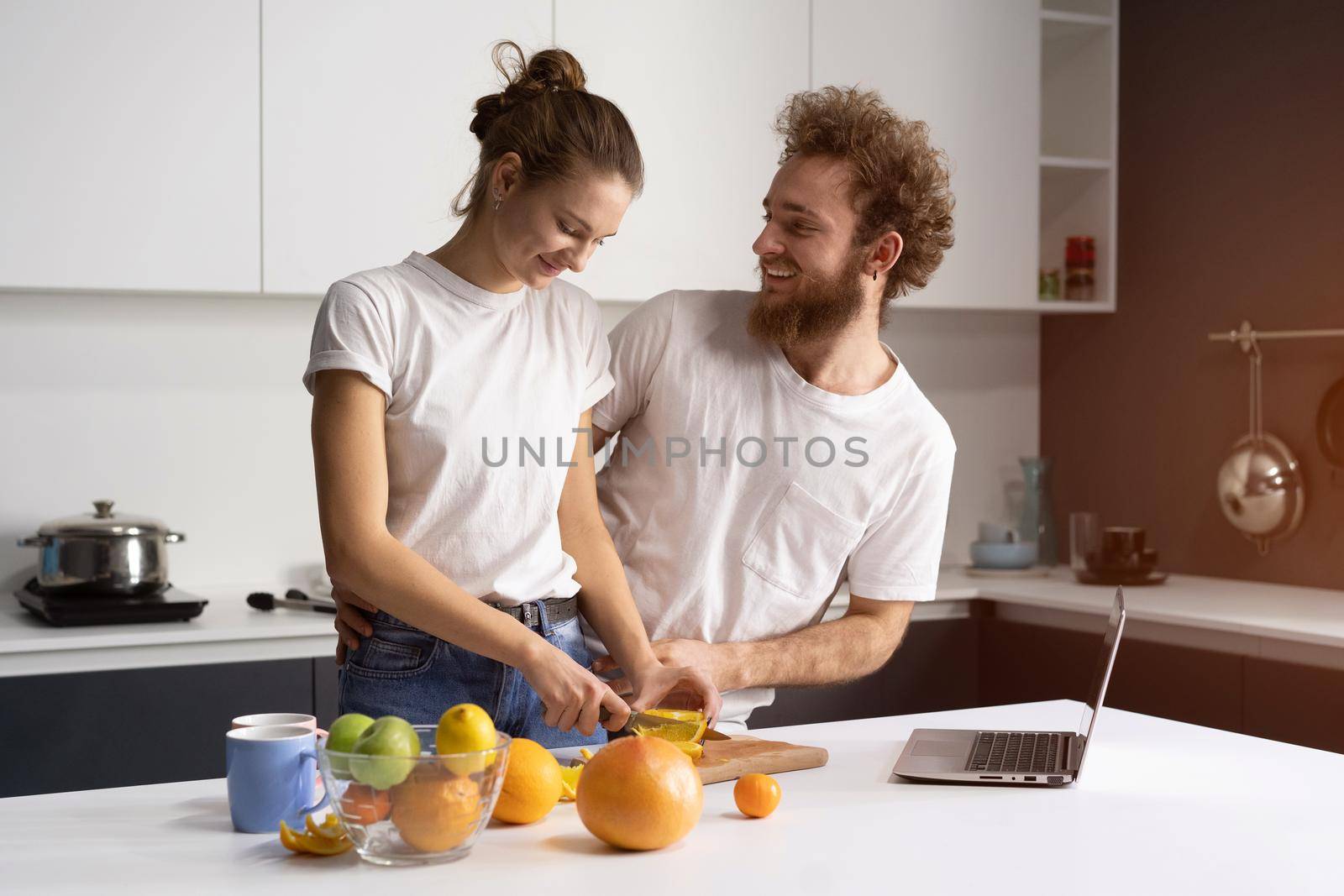Happy of their new home young couple cooking healthy food in new kitchen watching online streaming videos on laptop. Beautiful young couple talking on video call using laptop. New home concept.