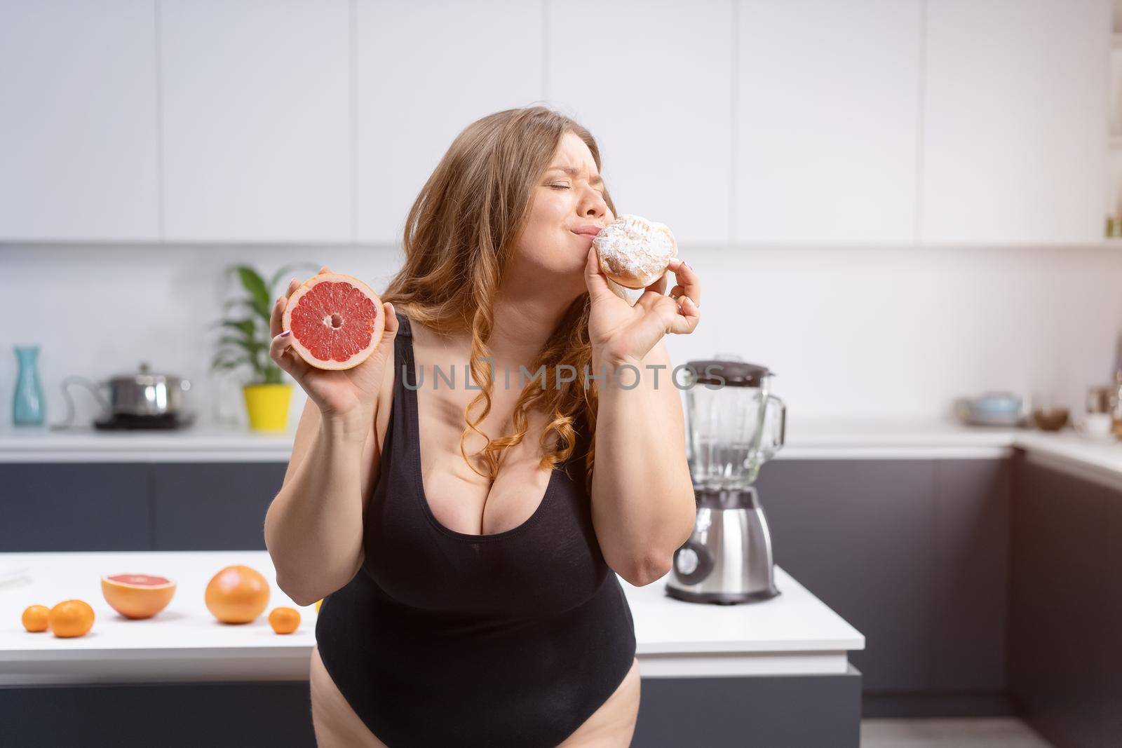 Choosing healthy or unhealthy food positive woman standing on the kitchen holding grapefruit and daughnut with lots fruits on table next to blander. Sexy overweight girl sitting on the kitchen table by LipikStockMedia