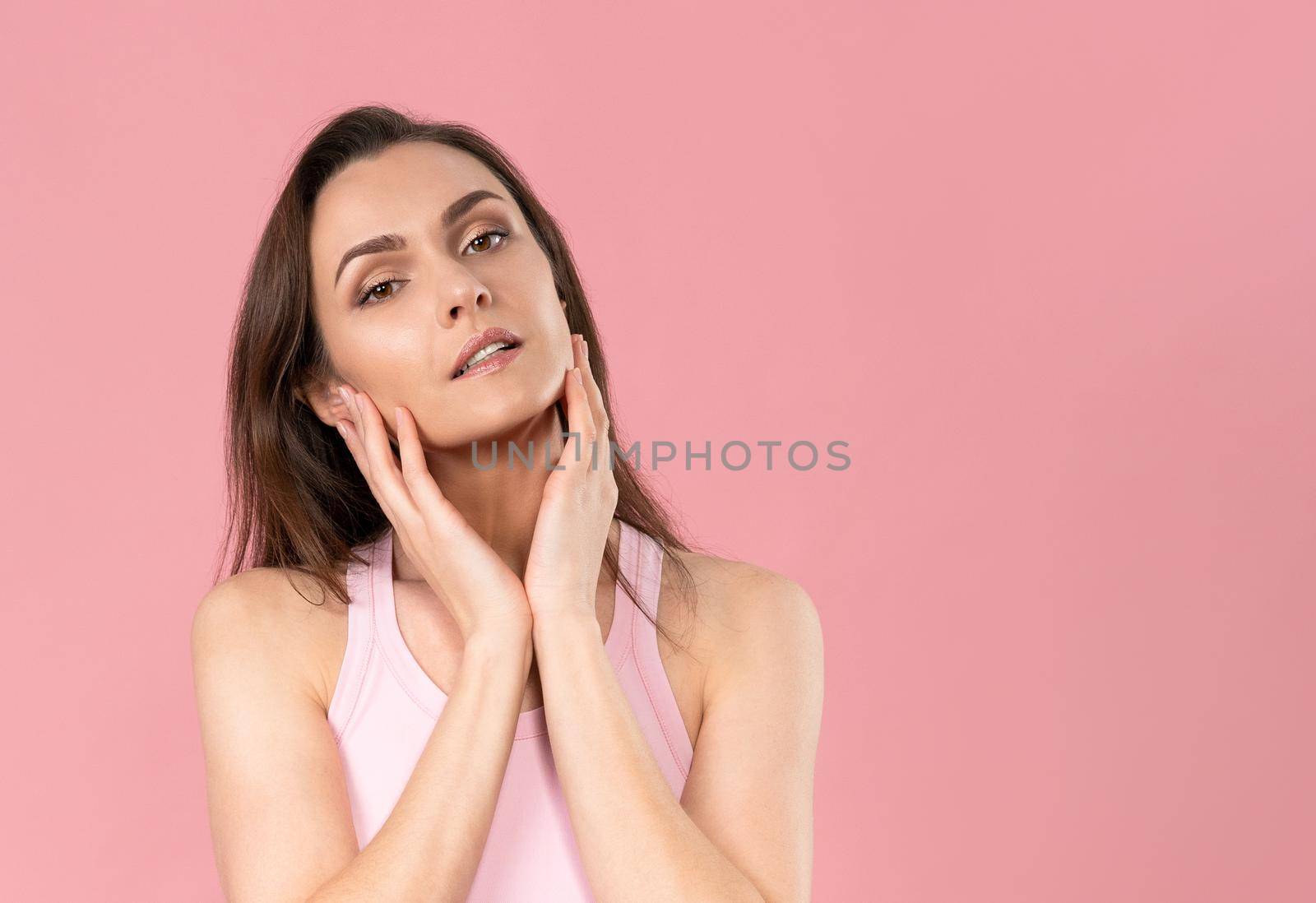 Moisturising her face beautiful woman with no makeup standing with hands on her chicks, massaging it gently attractive brunette girl on pink background. Skin care concept.