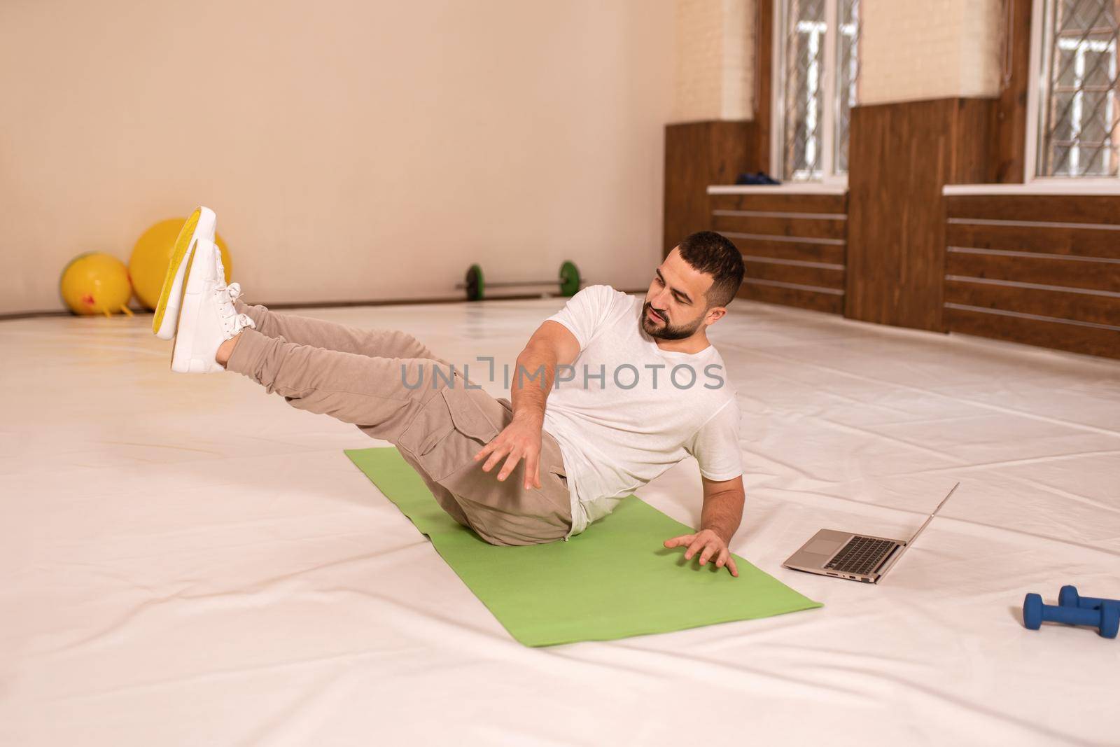 Sexy fitness trainer doing crunches sitting on the mat pad with laptop doing online video conference with a client during self isolation. Young man doing special exercises for abs muscles.