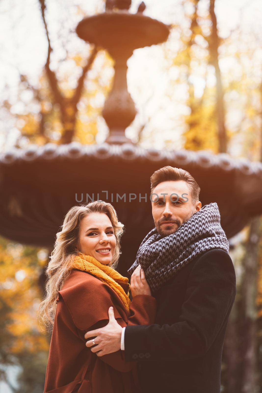 Half-length portrait loving couple of young people standing embracing and looking at camera with an old fountain on background at the autumn park wearing autumn coats. Toned photo.