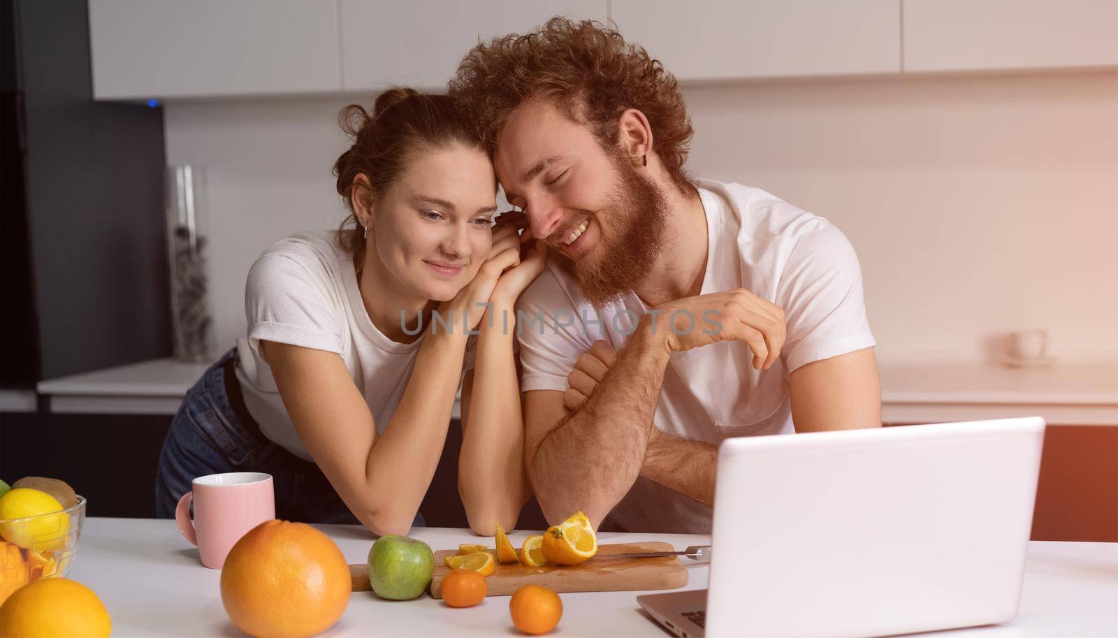 Beautiful young couple talking on video call using laptop. Young couple cooking healthy food in kitchen at home. Man leaned on girl smiling watching romantic movie by LipikStockMedia