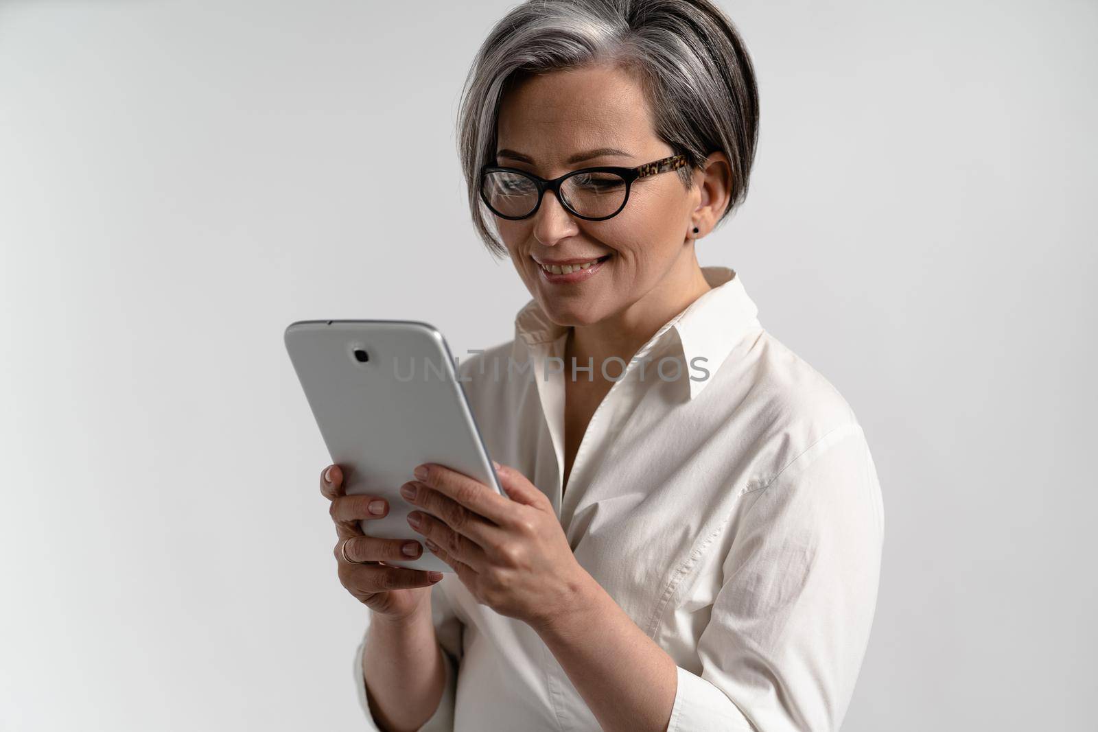 Portrait of a grey haired senior woman in white shirt and eye glasses using digital tablet. Technology by the elderly browsing internet shopping online using pad isolated on white background by LipikStockMedia