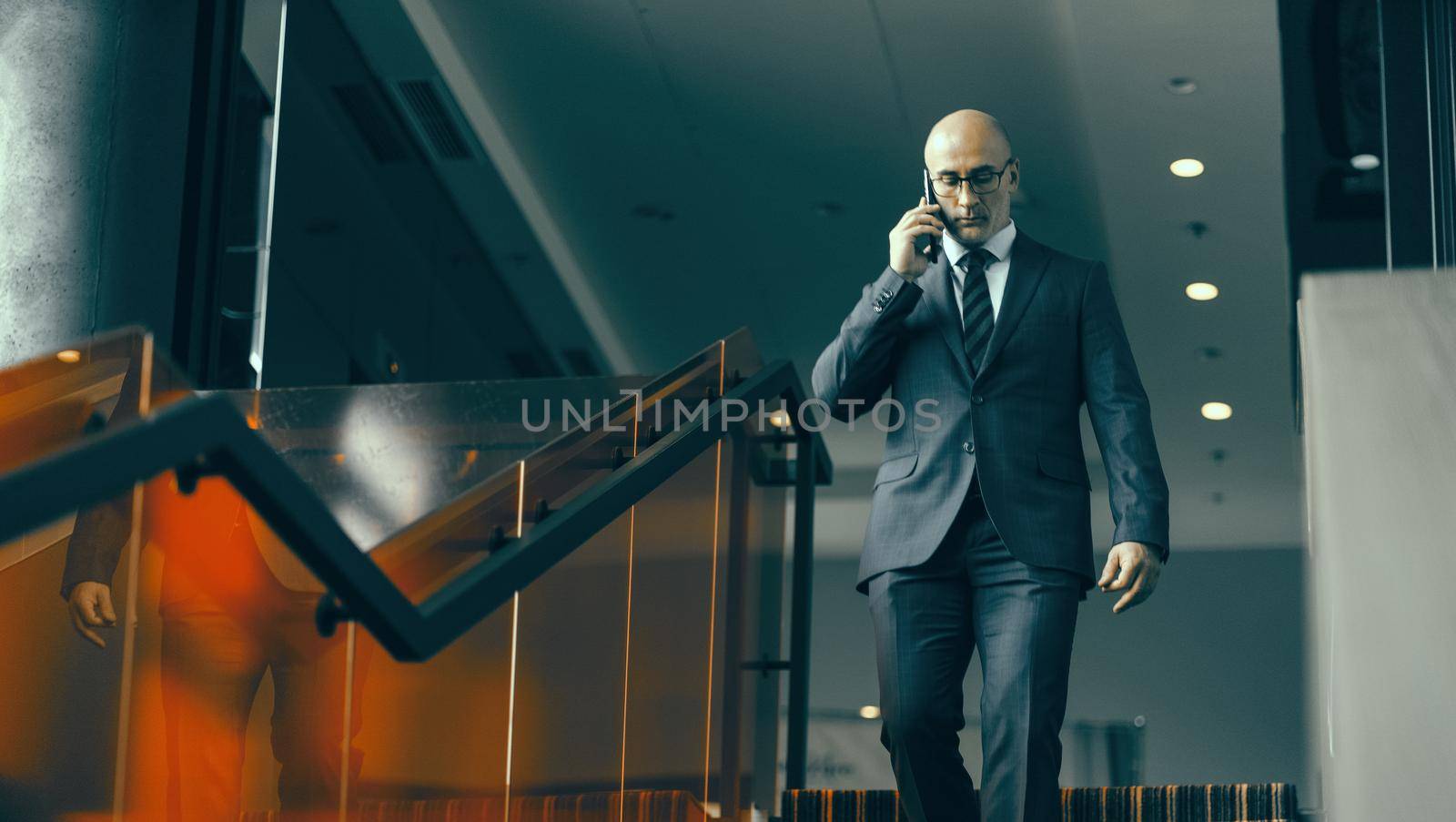 Walking down the stairs and having a conversation on the phone mature bald businessman. Handsome concerned man walking down modern office stairs by LipikStockMedia