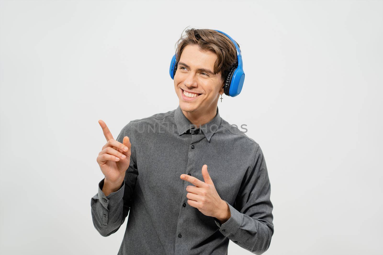 Happy smiling young guy in grey shirt standing listening music wearing blue wireless headphones pointing fingers sideways. Funny young guy listen to his favourite track or song.