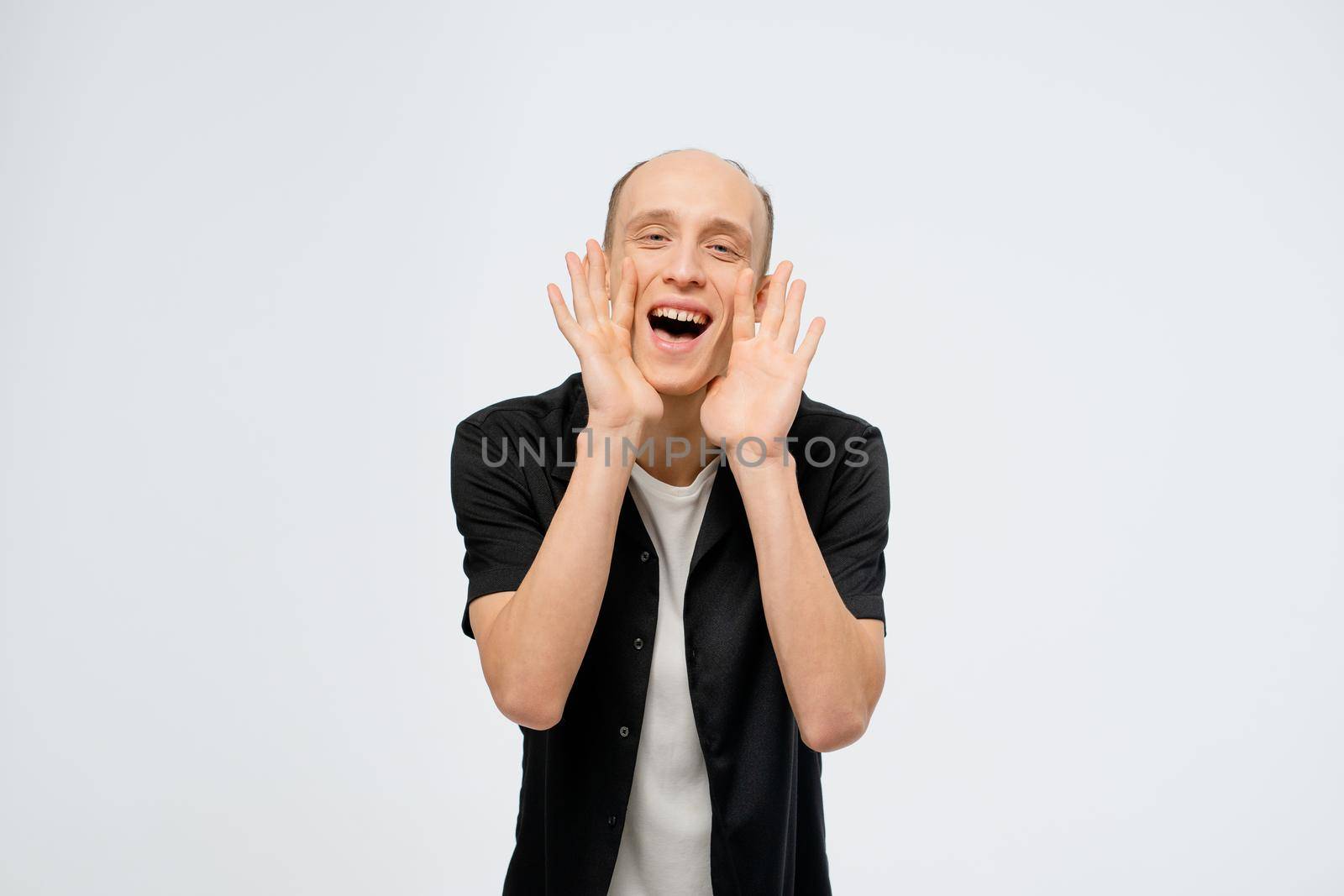 Screaming, delivering information young bald man wearing black shirt with white t-shirt underneath. Excited young adult happy man showing satisfaction with hands over white background in studio.