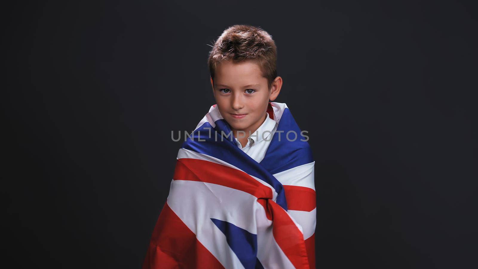 Little boy patriot standing side turned wrapped in a UK or British flag celebrates independence day expresses patriotism isolated on black background by LipikStockMedia