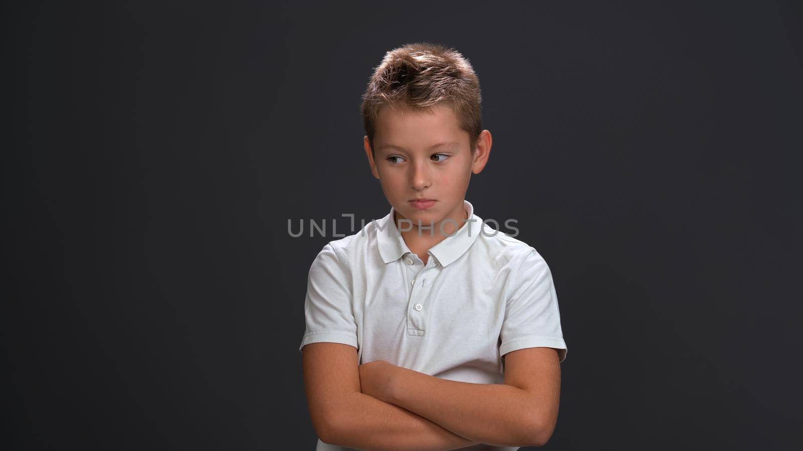 Unhappy or sad little boy looking a side frowning wearing white polo shirt and black pants isolated on black background.