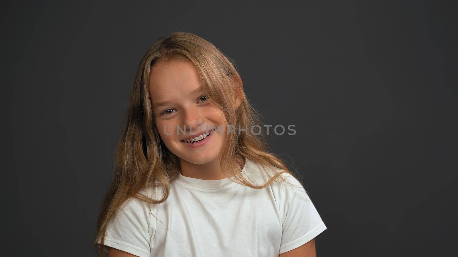 Smiling girl with bow sideways head looking at the camera wearing white t-shirt isolated on dark grey or black background. Child emotions concept.