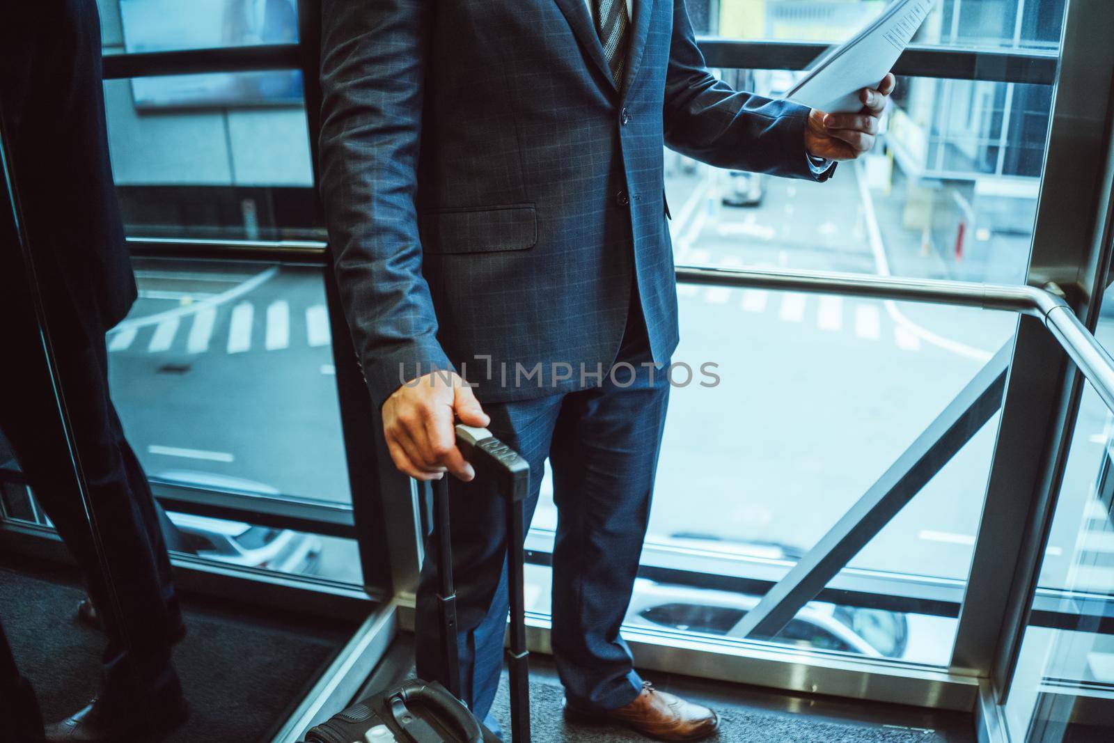 No face visible. Businessman looking at documents wearing a suit standing next to the window with suitcase next to him in the modern office with street view. Man holding a papers next to the window by LipikStockMedia