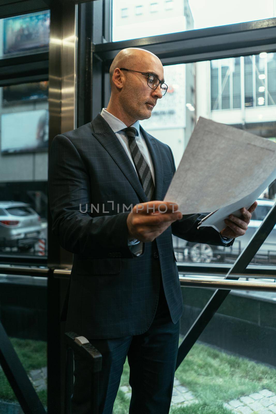 Business man looking at papers next to the window. Businessman looking at documents wearing a suit standing next to the window with suitcase next to him in the modern office with street view by LipikStockMedia