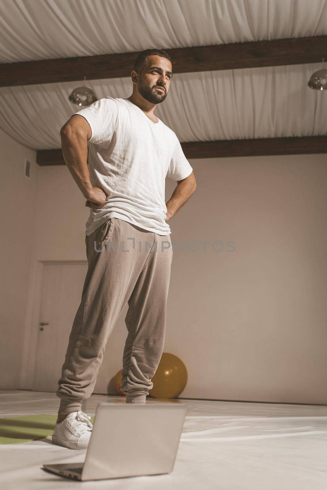 sportive young man trains hard at home standing in front of laptop. Self isolated and motivated young man warm-up doing special exercises for muscles. Work out during coronavirus lockdown by LipikStockMedia