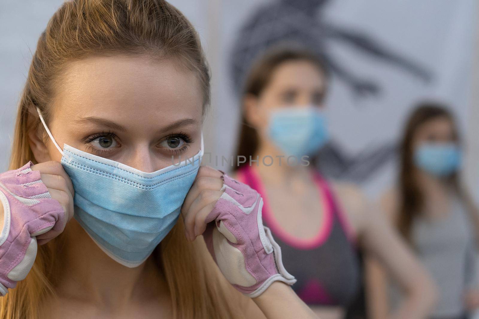 Putting on medical face protective mask athletic girls in sports out fits standing in yoga class, standing one after another in selective focus. Group of young sporty people practicing yoga lesson by LipikStockMedia