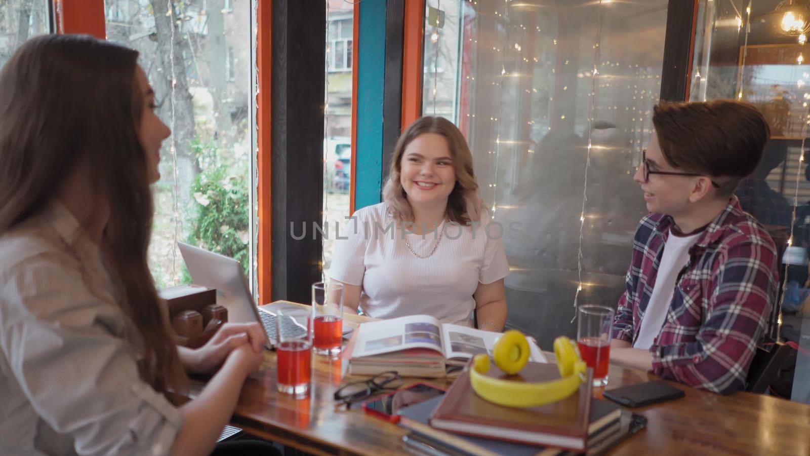 Three young students guy and girls study while having a brake in between lectures reading books and working on laptop having discussion sitting in cafe with lights running on background. 4K footage by LipikStockMedia