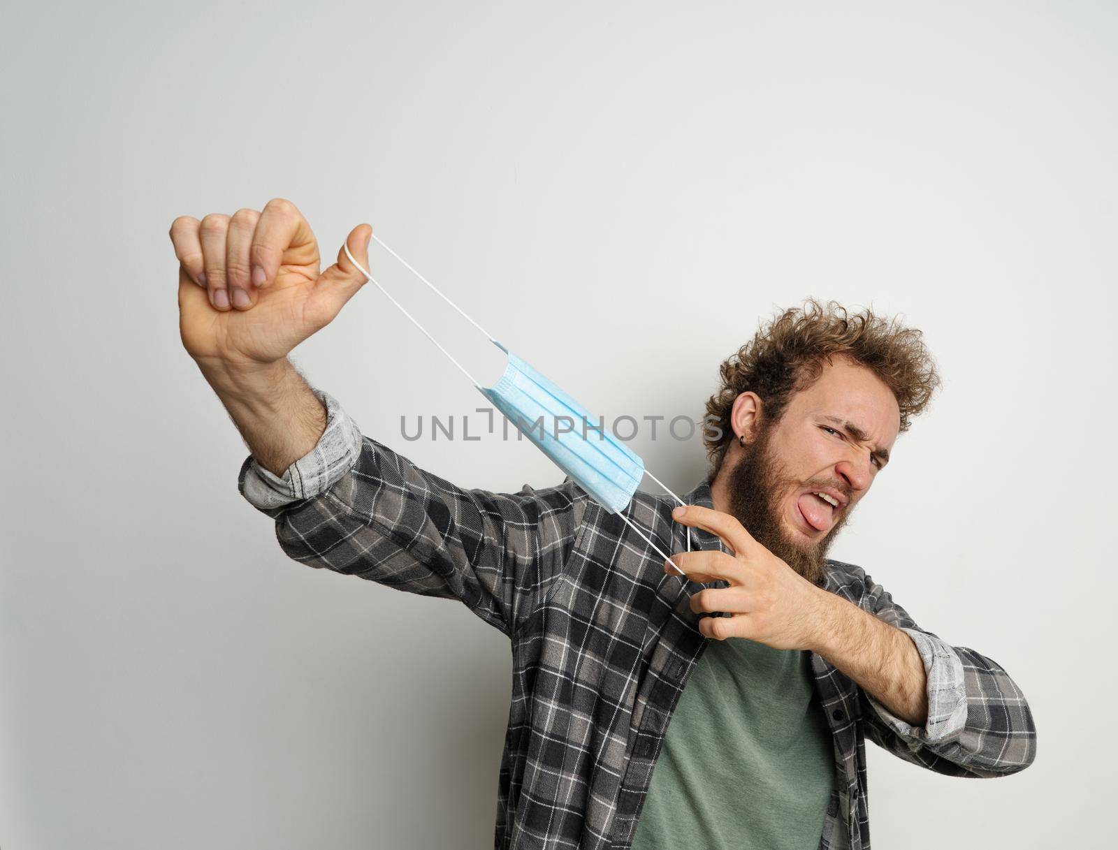 Played or got rid of young man holding protective medical mask in front of face ready to end quarantine, with curly hair, wearing plaid shirt and olive t-shirt under. White background by LipikStockMedia