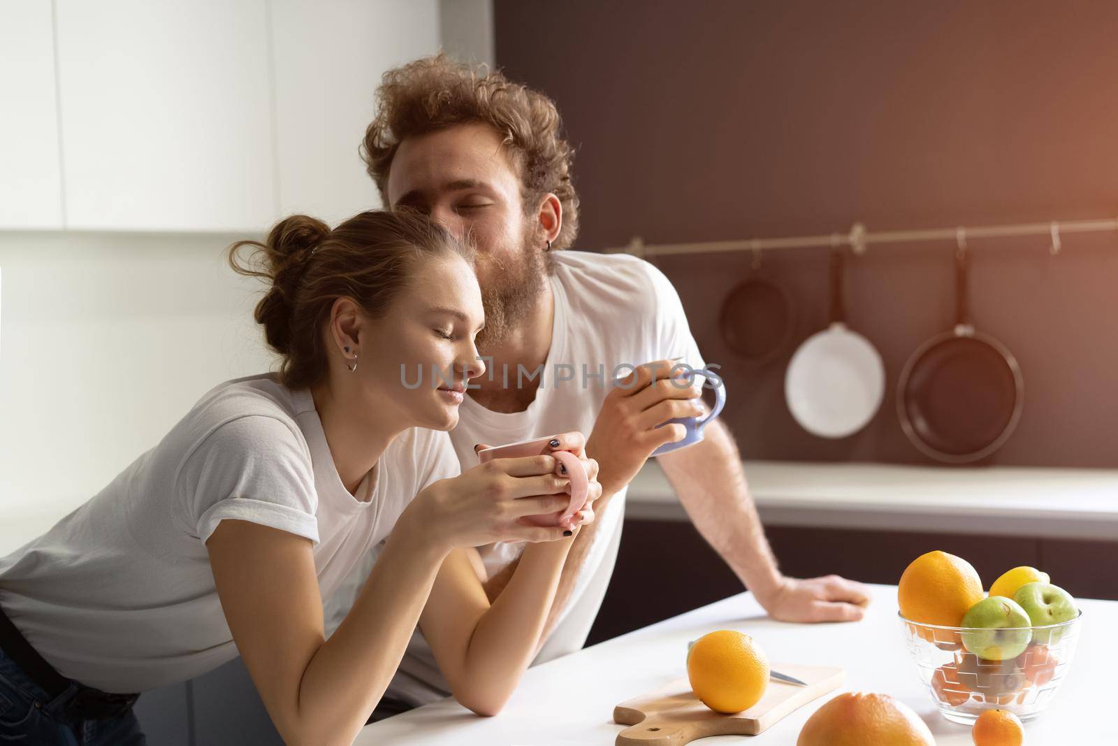 Morning coffee and gentle kissing feeding each other making fun at modern kitchen and smiling while cooking at home. Pretty girl feeds or nursing her boyfriend by LipikStockMedia