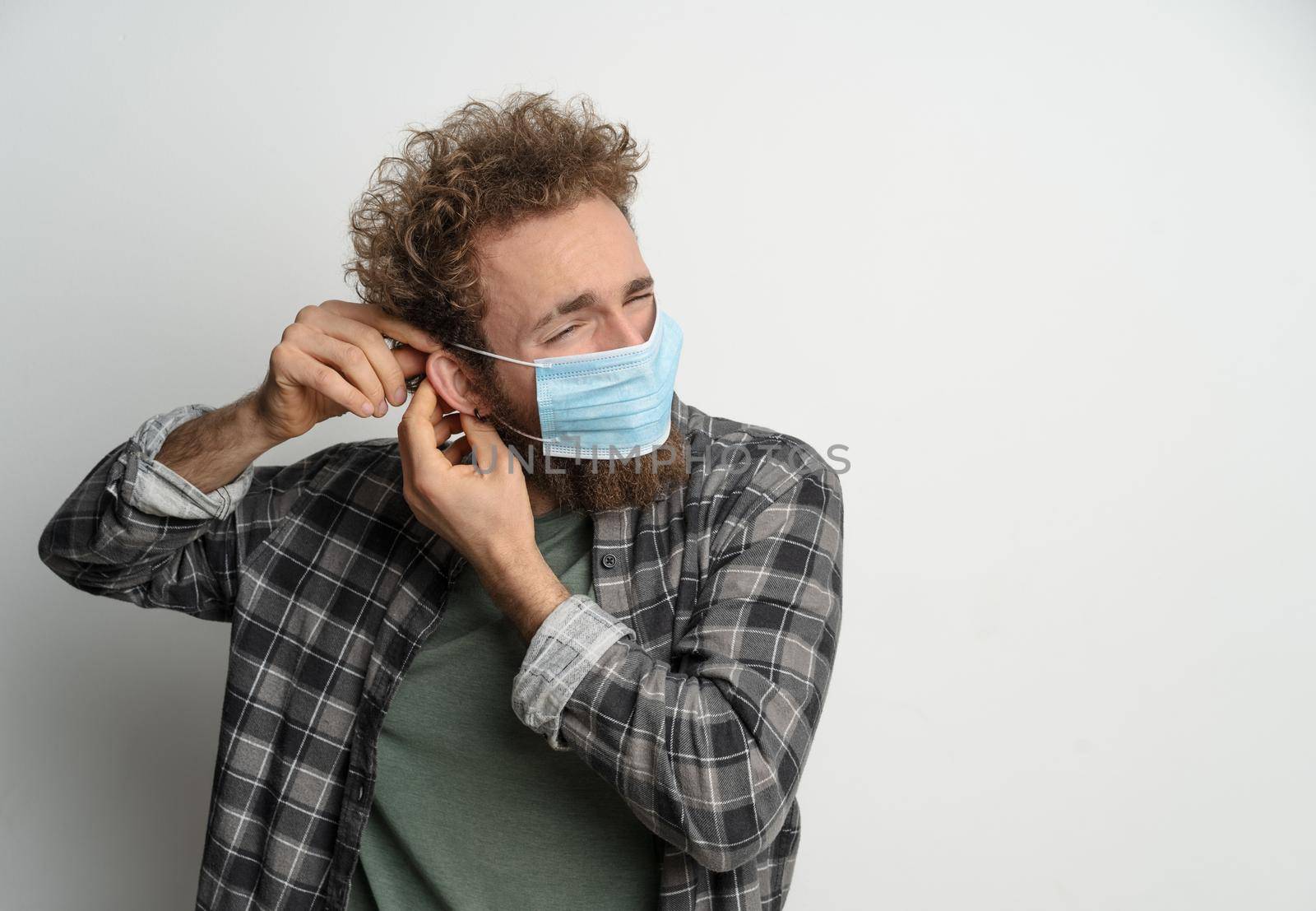 Putting on or taking off protective sterile medical mask on his face to protect coronavirus, with curly hair, young man wearing plaid shirt and olive t-shirt under. White background by LipikStockMedia