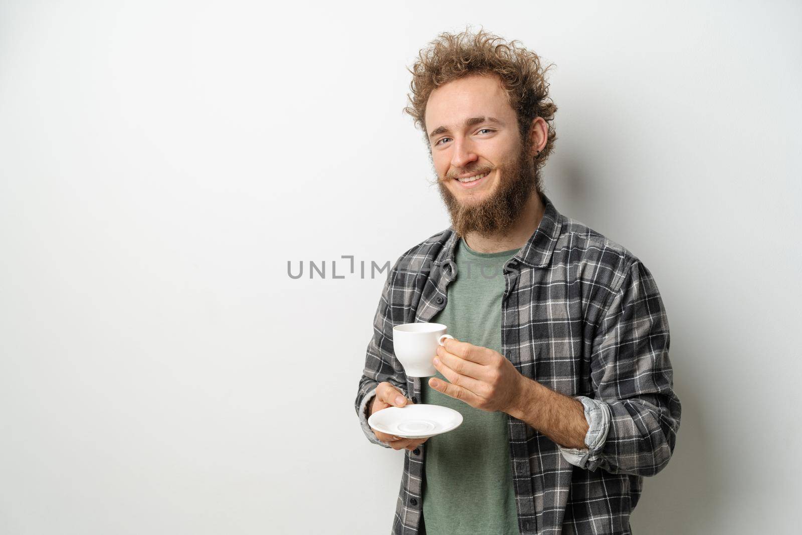 Smiling handsome man with curly hair and beard drinking coffee holding cup, wearing plaid long sleeve shirt isolated on white background.