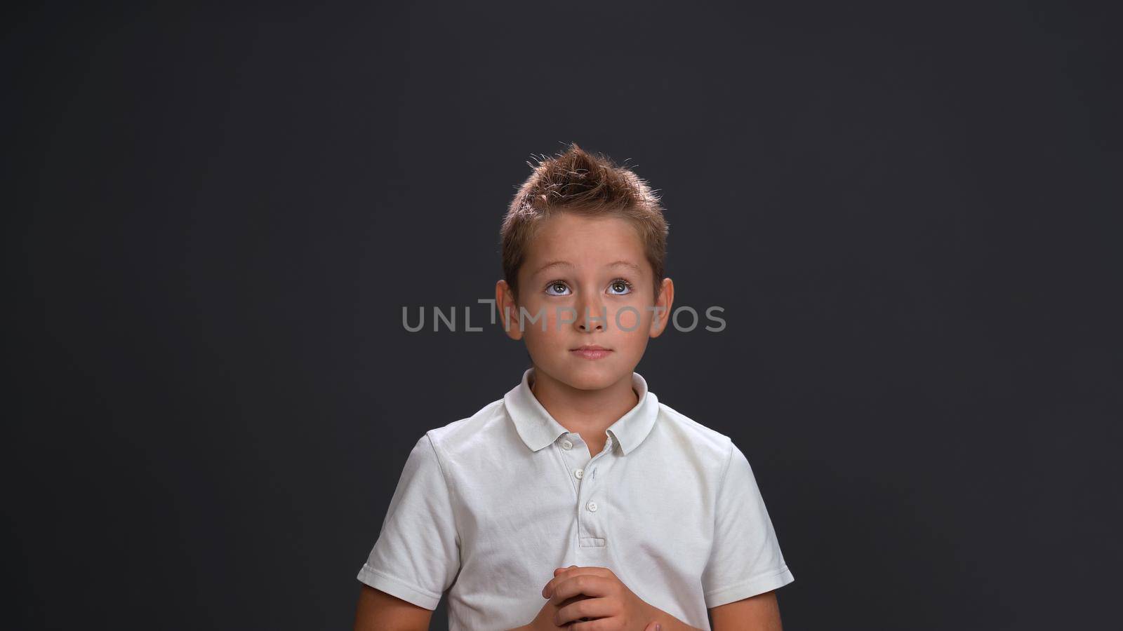 Expecting miracle boy in white shirt holding hands together in front of him while looking sideways at upper corner. Isolated on black background.