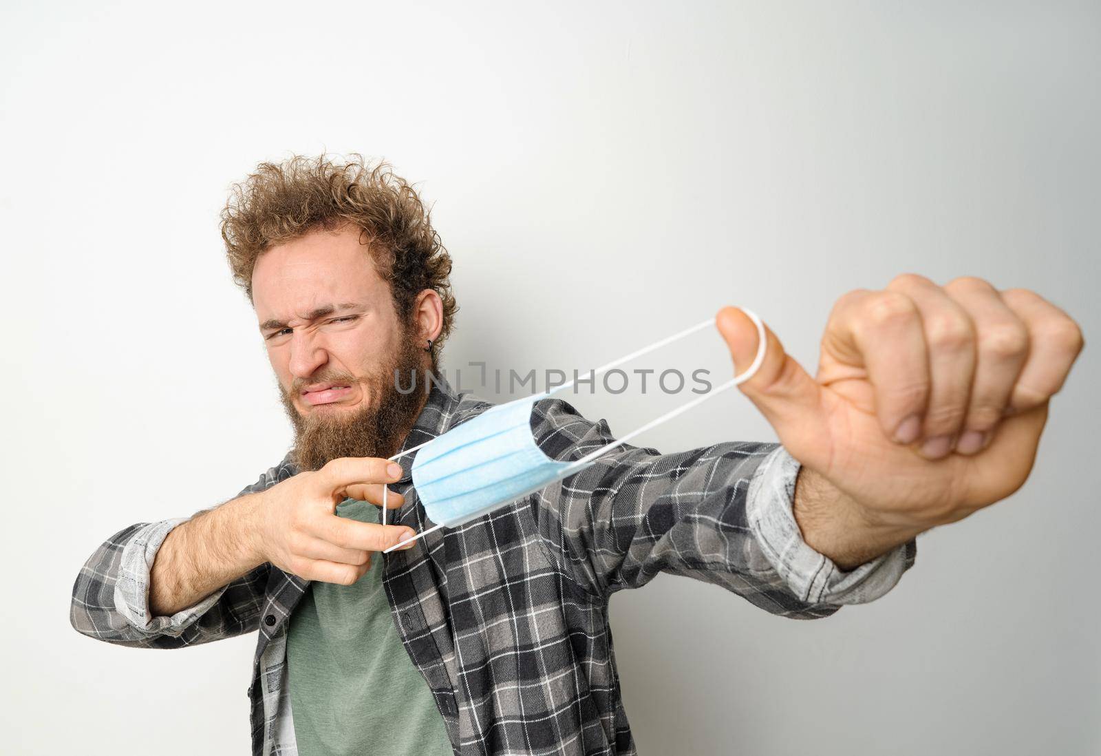 Getting rid of protective medical face mask young handsome man holding it stretching it in front of face wearing plaid shirt and olive t-shirt under. White background.