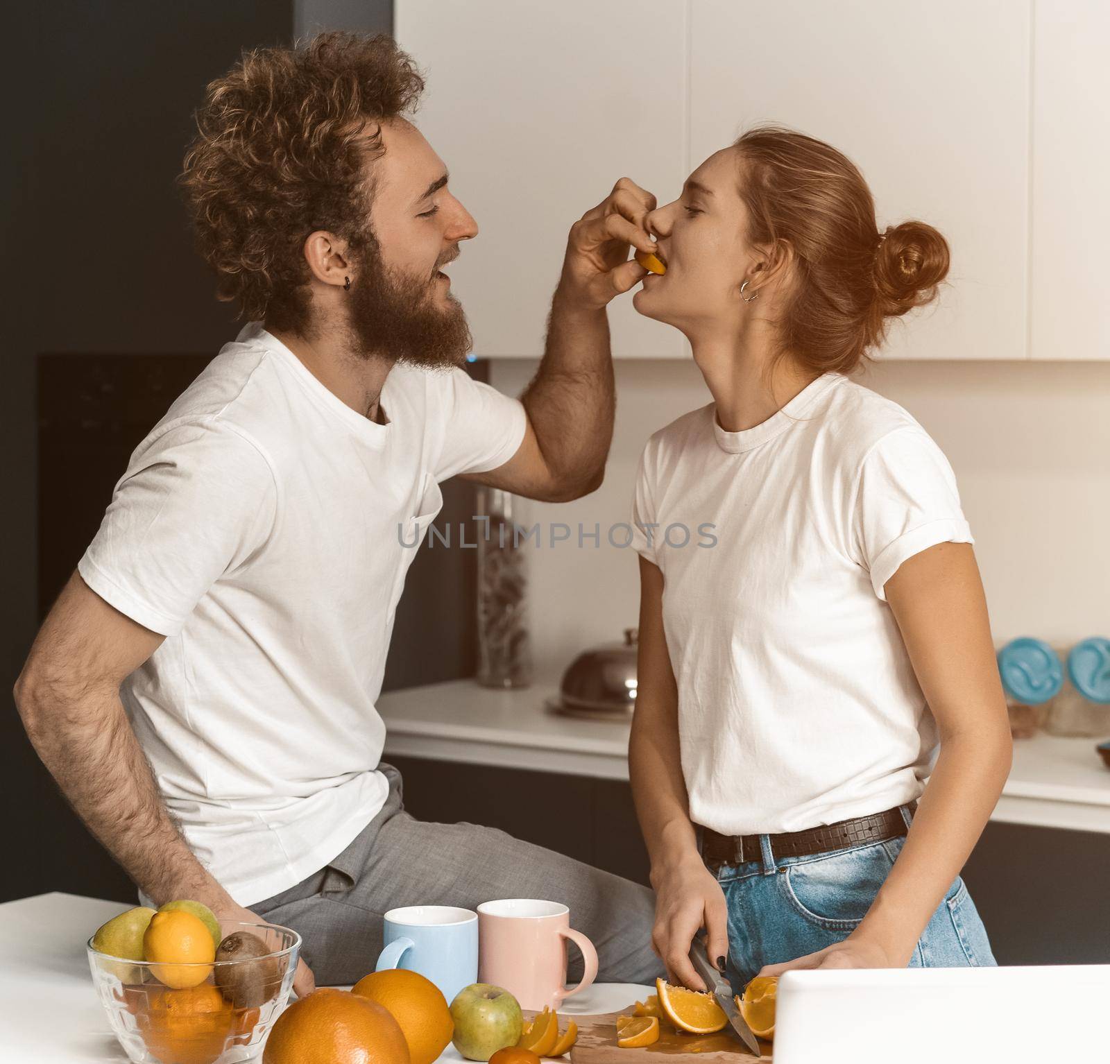 Boyfriend feeds or nursing his girlfriend. Beautiful young couple feeding each other making fun at modern kitchen and smiling while cooking at home by LipikStockMedia
